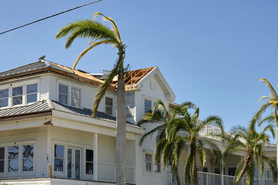 Storm-damaged roof on a white house with palm trees under a clear blue sky
